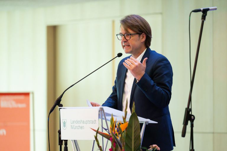 Stadtschulrat Florian Kraus steht an einem Pult mit Mikrofon seitlich stehen Blumen, Hinterlöpfe der Gäste im Vordergrung