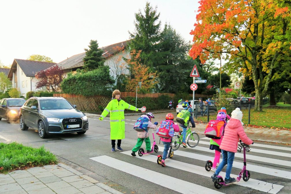 Schulweghelfer*in unterstützt am Zebrastreifen Kinder beim Überqueren der Straße