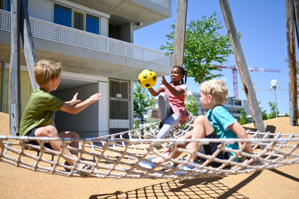 drei Kinder spielen mit einem Ball auf einem Seilnetz sitzend auf dem frei zugänglicher Pausenhof auf dem Gelände des neuen Bildungscampus Freiham