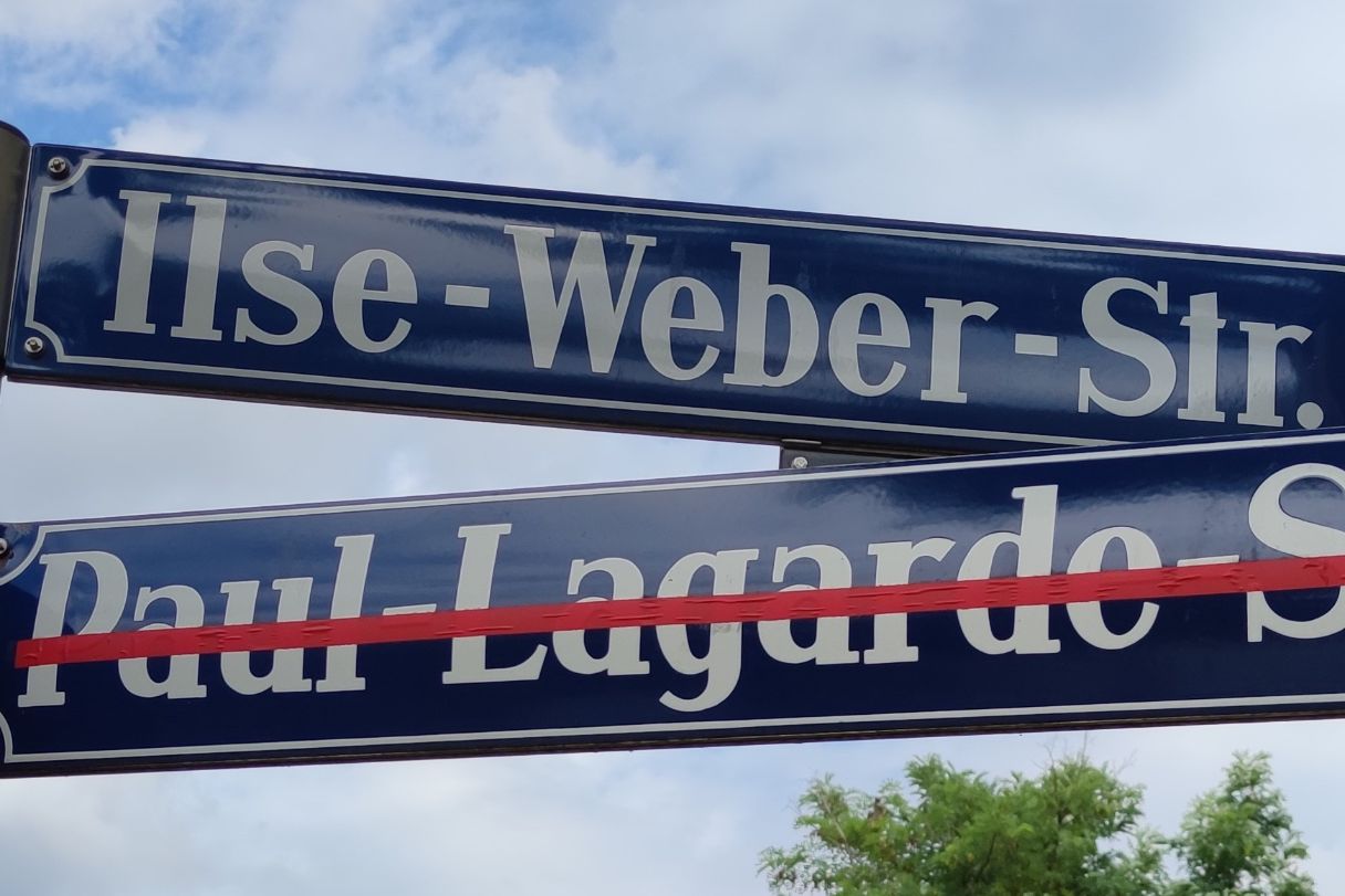 Das Bild zeigt zwei Straßennamenschilder übereinander an einem Pfosten: der Name Paul Lagard Straße wurde mit Klebeband durchgestrichen, der Name Else Weber Straße ist neu hinzugekommen.