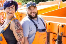 Zwei lachende Menschen stehen vor orangenen Abfallcontainern