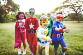 Auf dem Bild sind zwei Mädchen und drei Jungs in bunten Superheldenkostümen zu sehen, die farbige Brillen tragen und auf einer Wiese auf den Fotografen zulaufen.