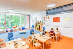 Gruppenspielraum des Haus für Kinder Wackersbergerstr. 71  mit Spielteppich, Kindertischen und -stühlen vor großer Fensterfront