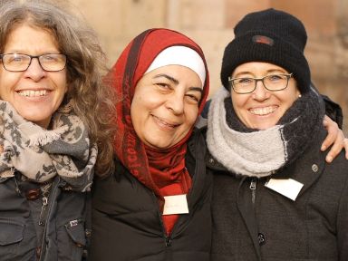 Drei Frauen umarmen sich und lächeln in die Kamera, eine davon trägt Kopftuch