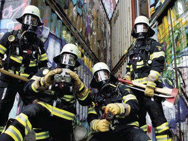 Vier Feuerwehrleute vor einem Graffitti, sie haben verschiedene Gerätschaften dabei