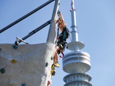 Ein Kletterer hängt vor dem Olympiaturm an der mobilen Kletterwand.