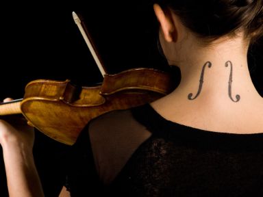 Eine Frau spielt Geige und ist mit dem Rücken zugewandt. Auf ihrem Nacken hat sie ein Tattoo in Form von zwei Schalllöchern. 