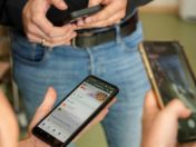Bild: Drei Personen bedienen ihre Smartphones. Im Vordergrund ist ein Smartphone mit der geöffneten App „KITAMuc by KIKOM“ zu sehen, auf der unter anderem aktuelle Mitteilungen und Termine angezeigt werden. Die Szene vermittelt einen Einblick in den digitalen Alltag in einer Kindertageseinrichtung.