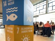 Zwei Würfel die SDG 14 und SDG 7 symbolisieren, dahinter Besucher im Munich Urban Colab